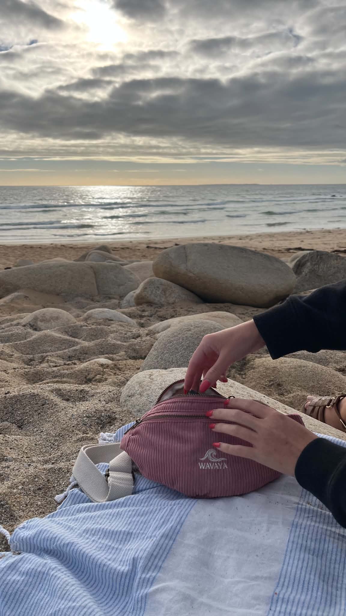 Sac banane rose en velours Wavaya posé sur une serviette à la plage face à l’océan
